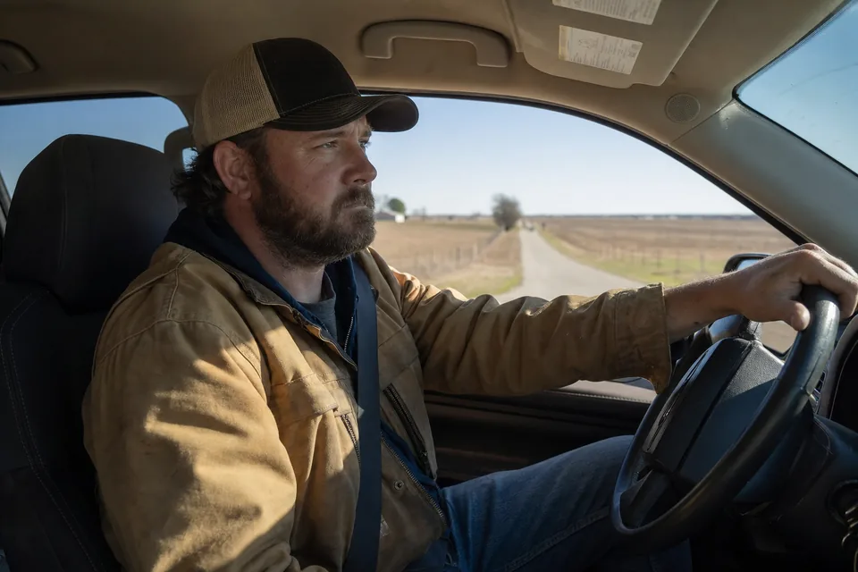 A rep driving a back road past open farmland, cap on, one hand on the wheel