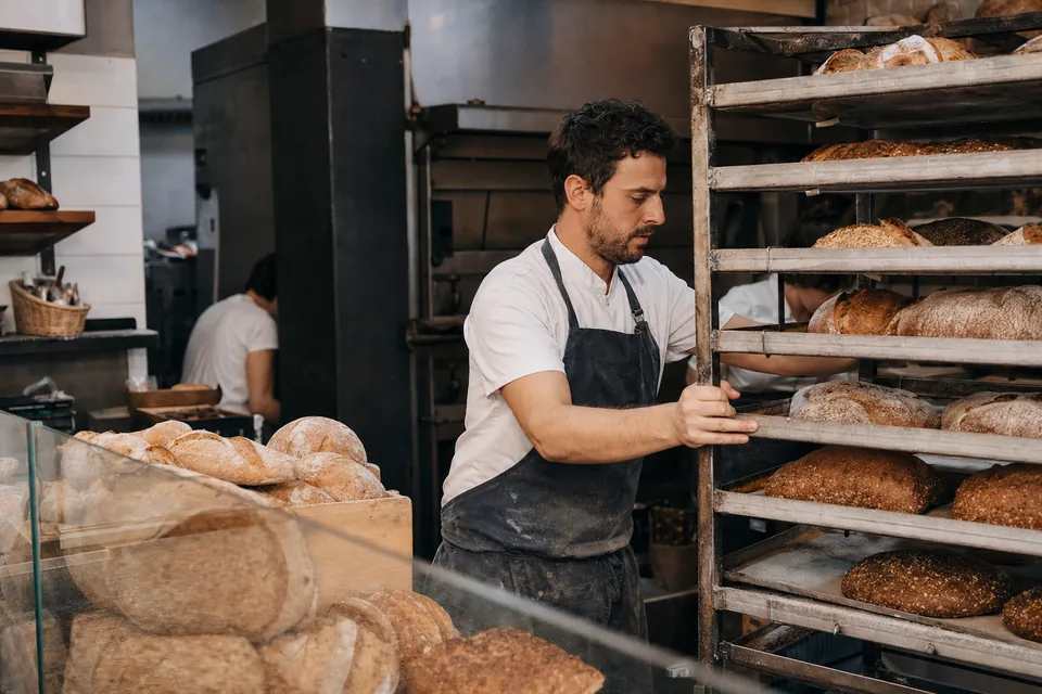 A baker pulling a rack of loaves past the front case of a bakery