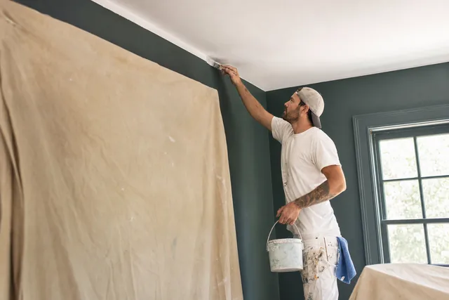 A painter cutting in a ceiling line above a drop cloth, paint can in hand