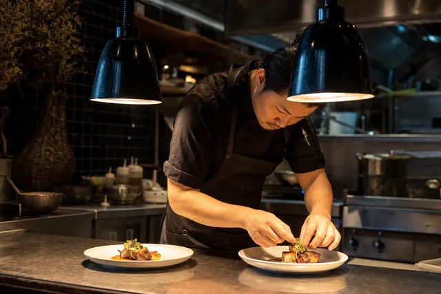 A chef plating two dishes under the pass lights on a restaurant line